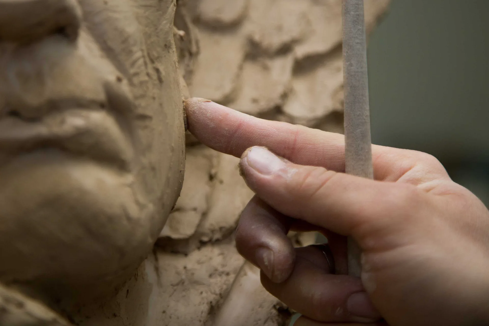 Close-up photograph of a hand touching carved stone or clay relief sculpture with classical figurative details.