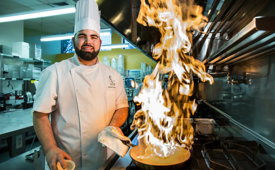 Chef in white coat performs high-heat cooking with dramatic flames rising from a pan in a professional kitchen.