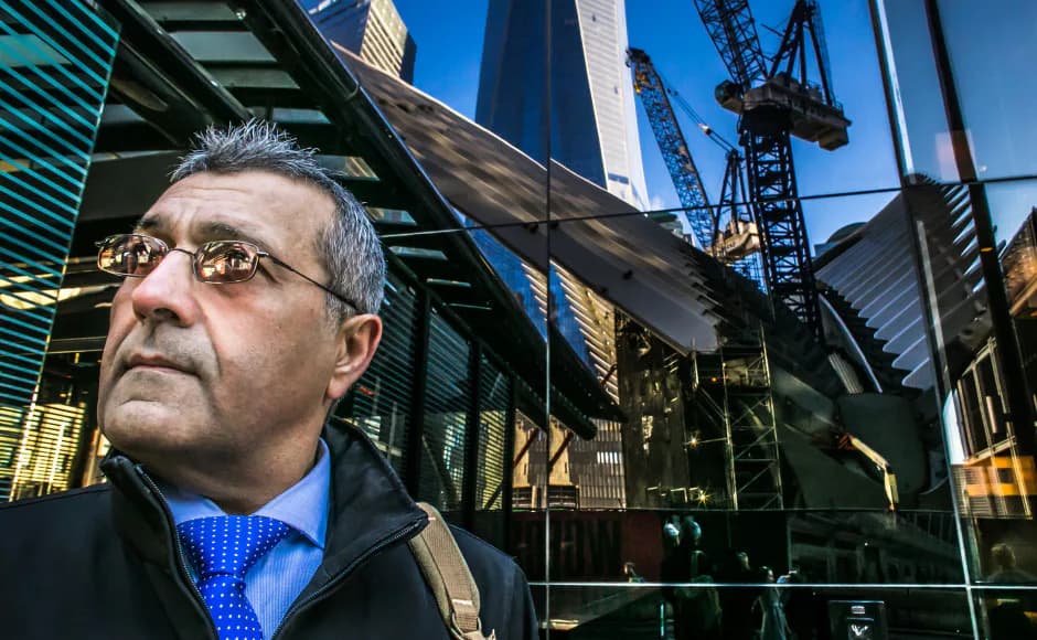 Middle-aged man wearing glasses, blue tie, and dark jacket photographed outdoors with construction cranes visible in background.