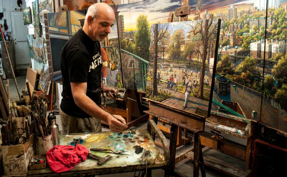 Artist in black shirt works at an easel with paintings of urban landscapes visible on the wall behind him.