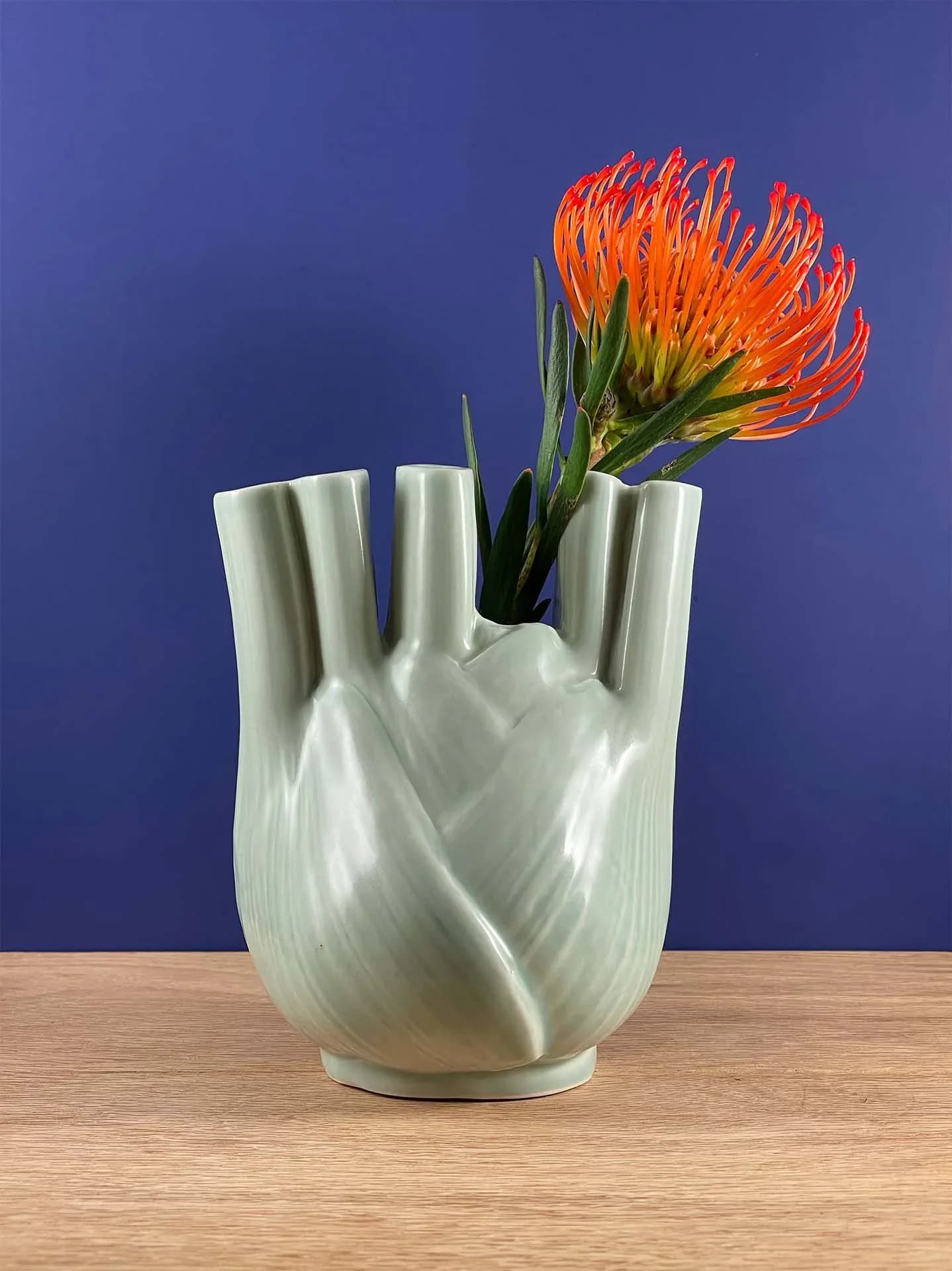 Celadon glazed fennel-form ceramic vase with orange protea flower on blue background