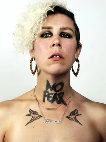Close-up portrait of a person with curly blonde hair, dramatic dark eye makeup, hoop earrings, and a 'NO FEAR' tattoo across their neck