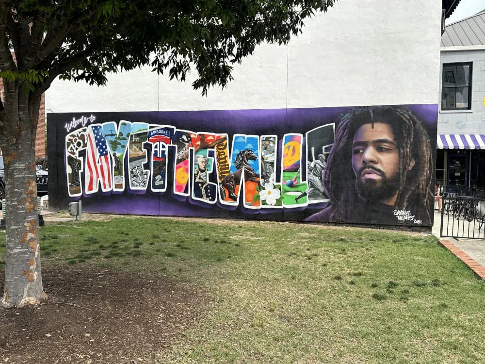Large-scale mural with purple background featuring colorful graffiti-style letters spelling a name, with a portrait of a man with dreadlocks on the right side.