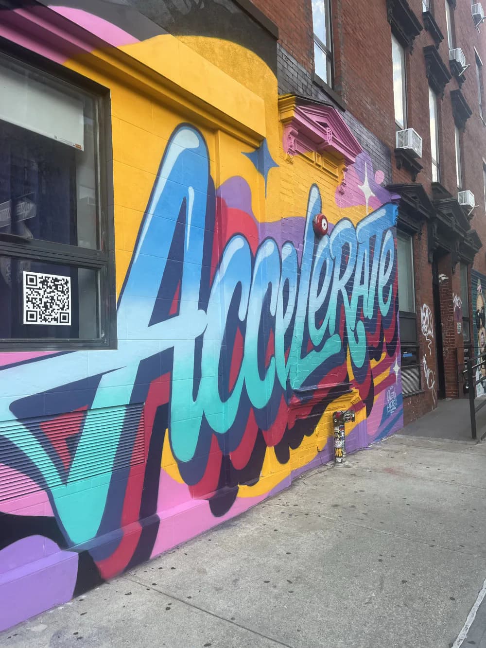 Vibrant street mural featuring large blue and cyan lettering on yellow, pink, and purple background with geometric shapes and a QR code on the left.