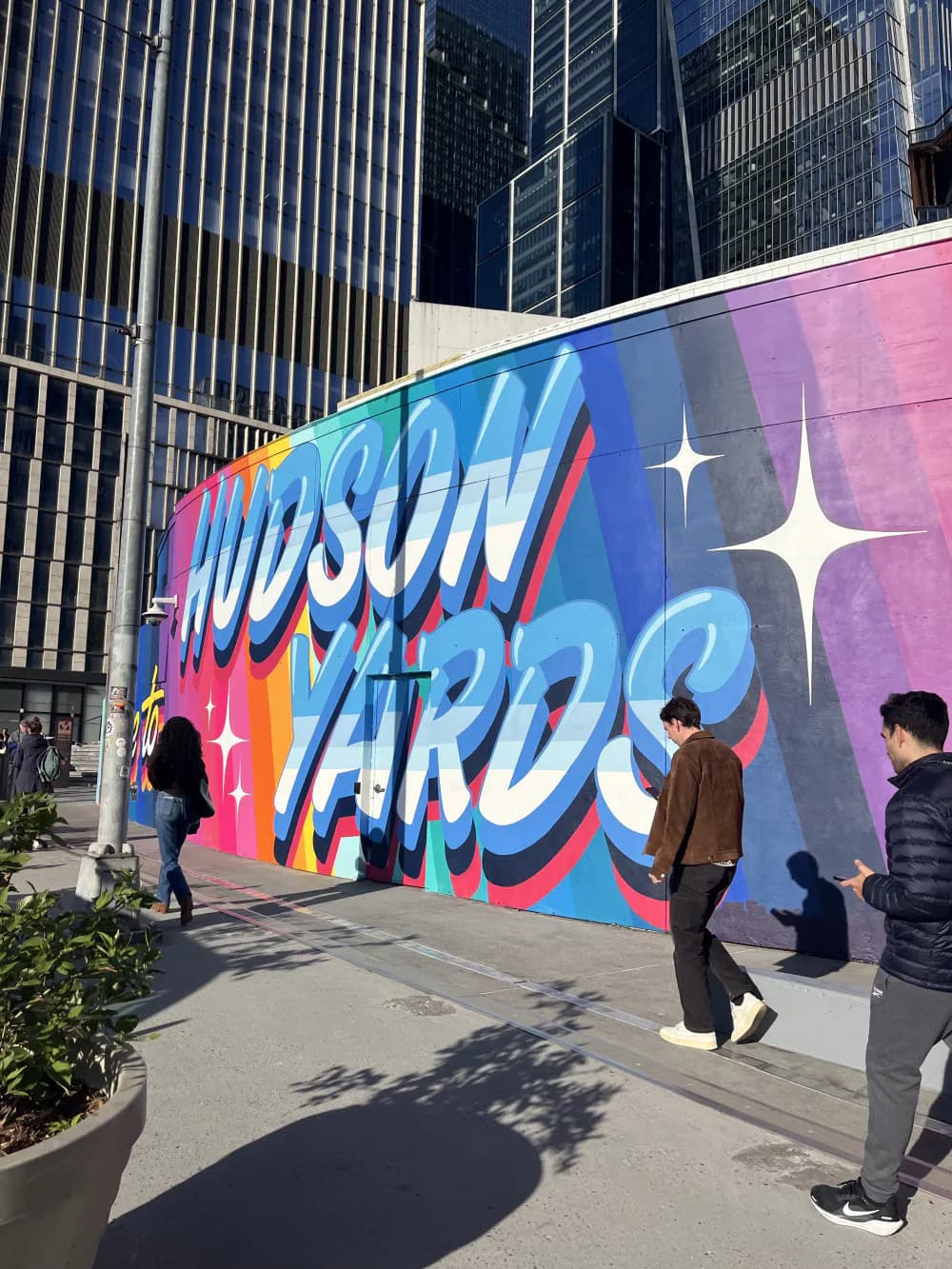 Large-scale mural with bold 'HUDSON YARDS' lettering in blue, pink, and orange on purple gradient background with white star accents, urban setting with pedestrians.