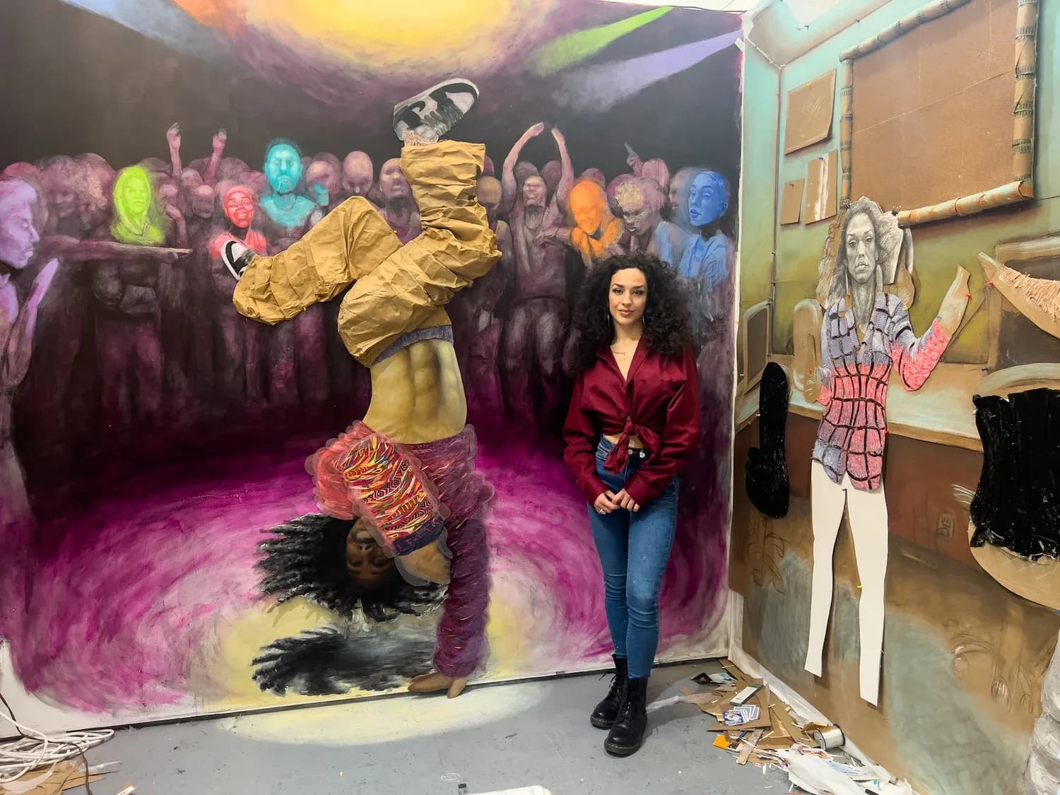 Mixed-media installation by Annie Legnini: 3D figure in golden-yellow clothing leaping above magenta swirls, with colorful crowd in background and life-size cutout figure on right.