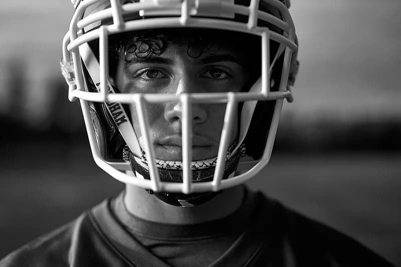 Black and white portrait photograph of a young man wearing a white football helmet with facemask, focused frontal composition against blurred background.