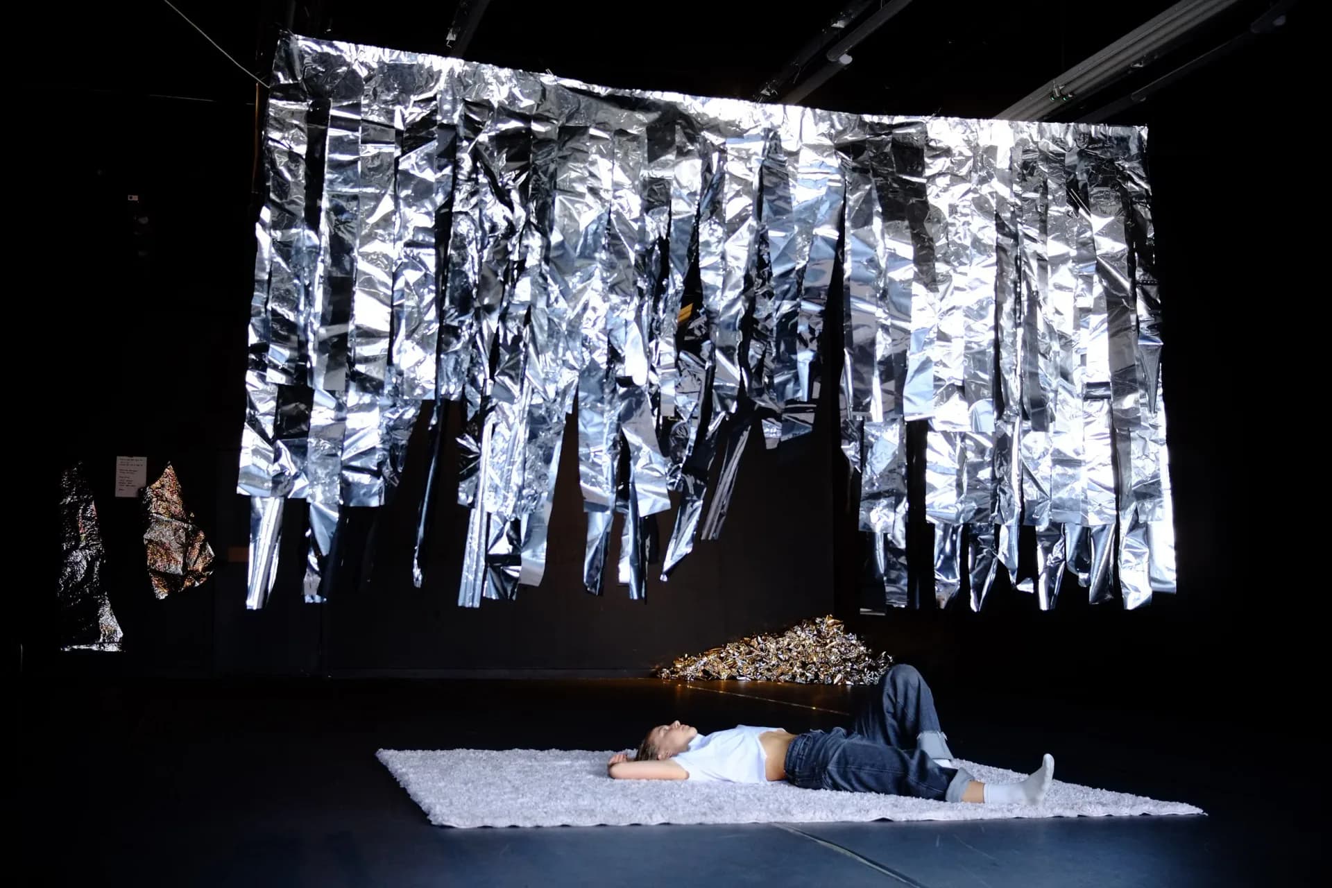 Performer lying beneath large suspended installation of silver mylar strips in a darkened theater space with a pile of crumpled foil nearby
