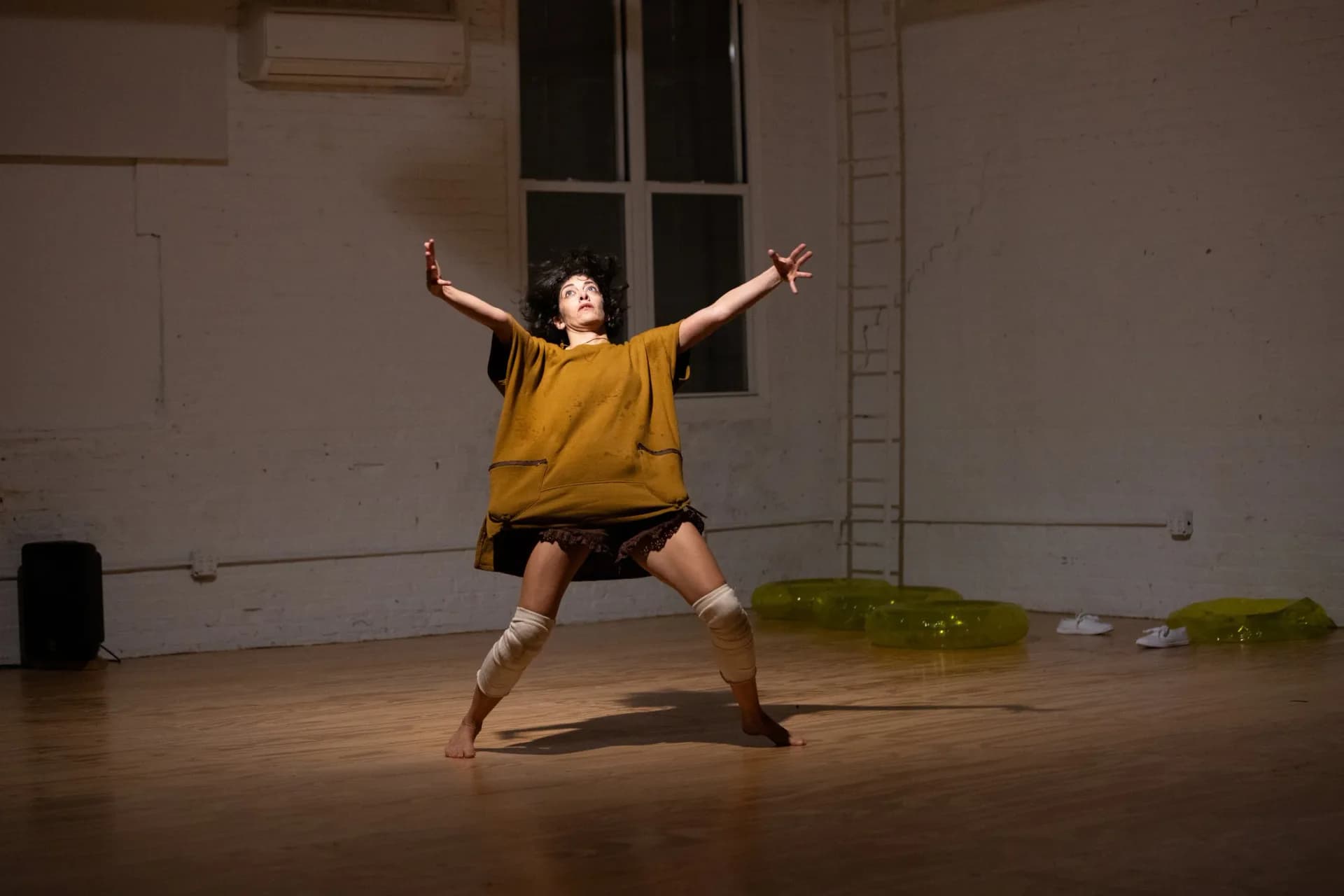 Performer in dynamic wide-stance pose with arms outstretched in a warmly lit studio space with yellow inflatable rings in the background
