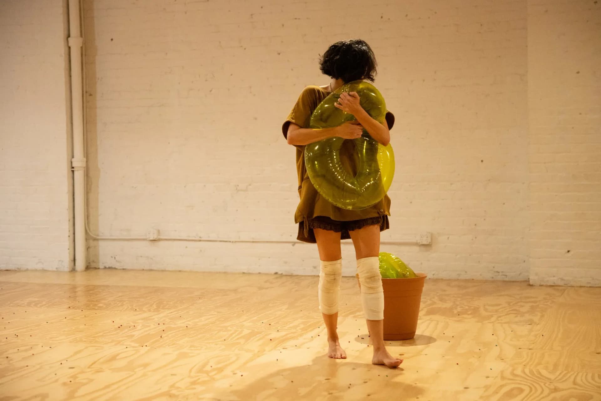Performer embracing a large translucent yellow inflatable ring with a terracotta pot containing another inflatable nearby on a plywood floor scattered with colorful pins