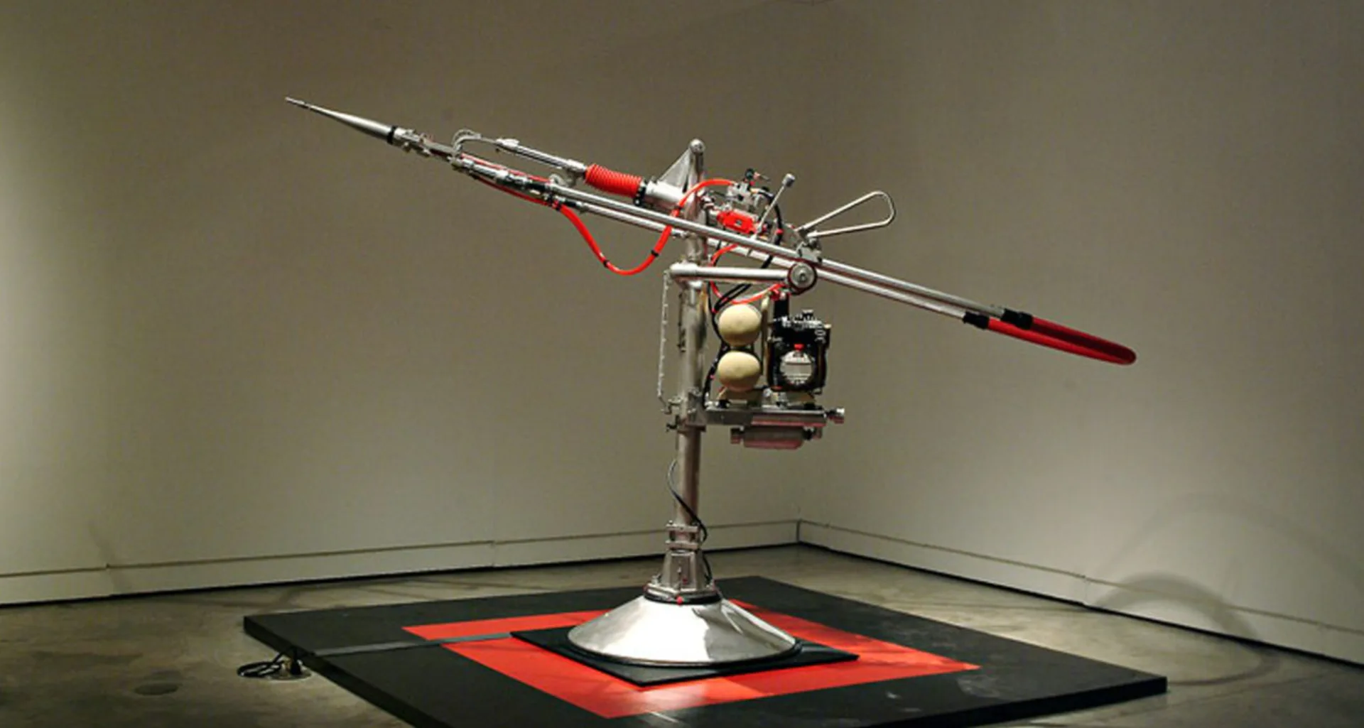 Kinetic sculpture by Arnaldo Morales with red metal base, rotating horizontal arms with weights, suspended within gallery space.