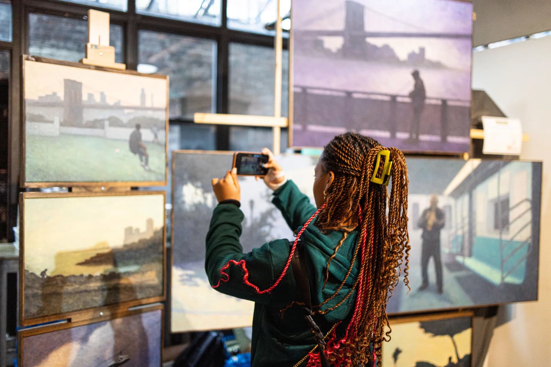 Person with braided hair photographing artwork displayed on gallery walls with Brooklyn bridge visible through large windows.