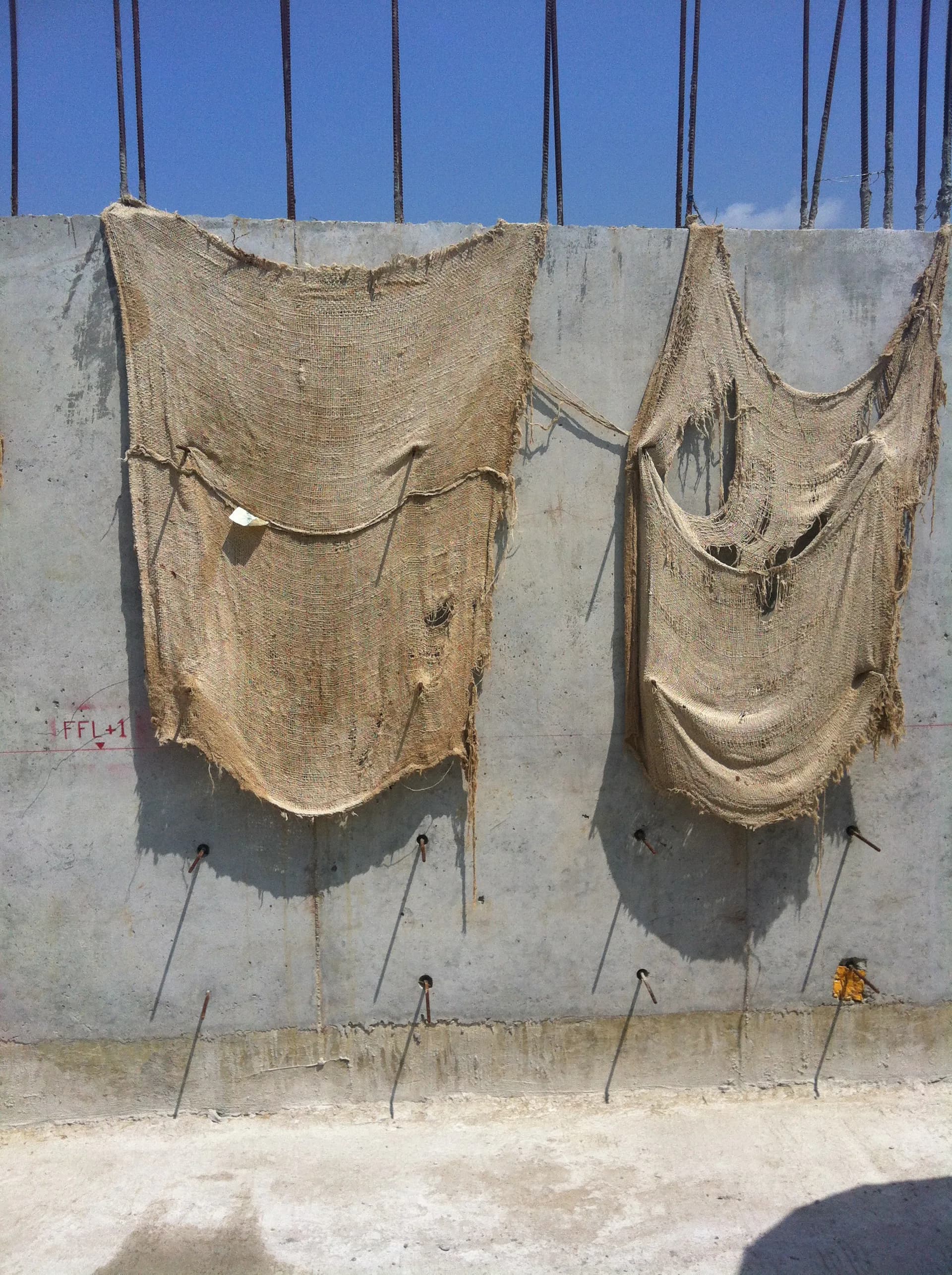 Photograph of two large woven burlap or textile panels hanging on a concrete wall against blue sky, casting shadows.