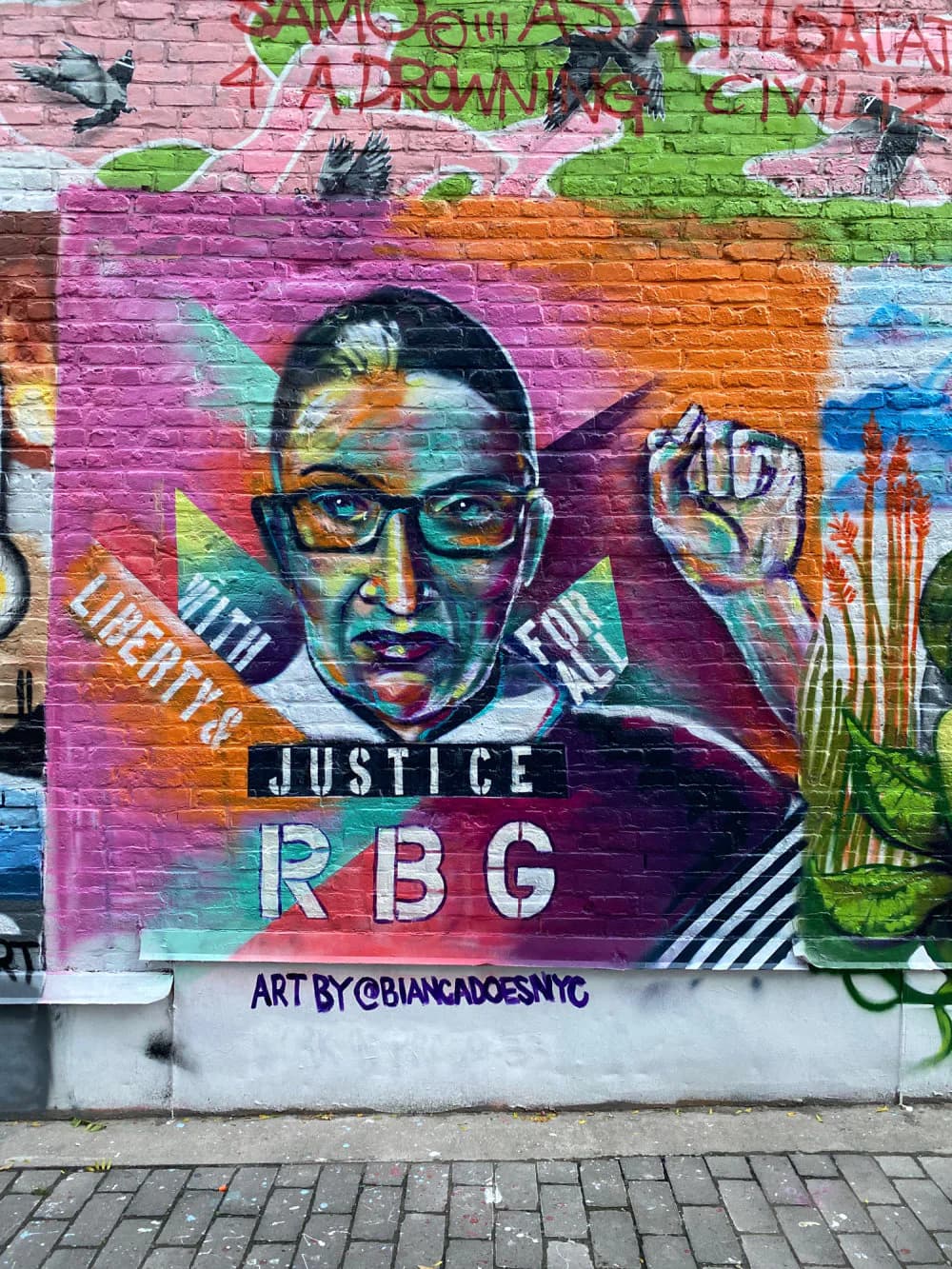 Vibrant street mural of a person wearing glasses with 'JUSTICE' and 'RBG' text, surrounded by colorful geometric shapes and green foliage elements.