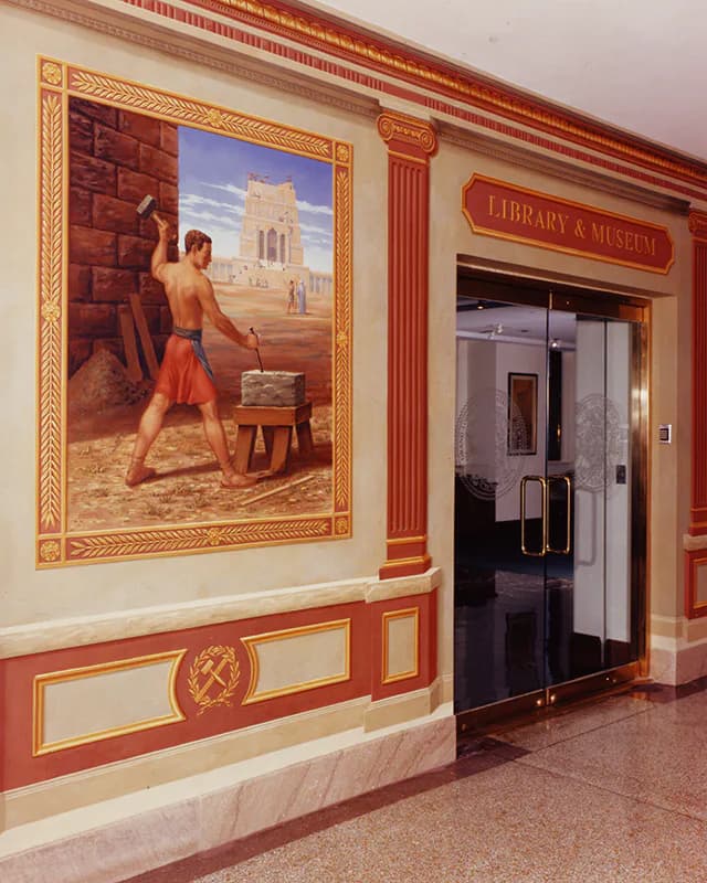 Museum or library interior hallway with ornate red and gold trim, framed mural artwork of a shirtless figure, and glass doors labeled 'Library & Museum'.