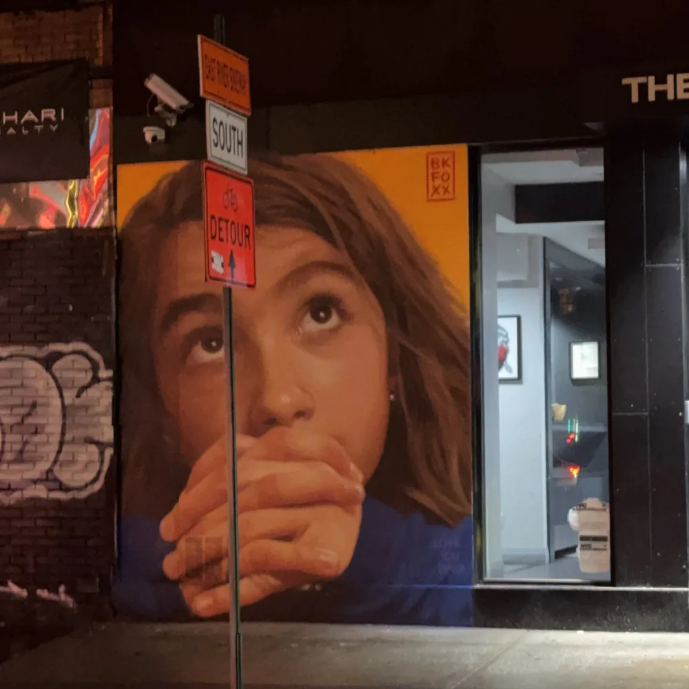 Mural of a woman's face with contemplative expression, rendered in warm tones and blue, on an urban storefront.