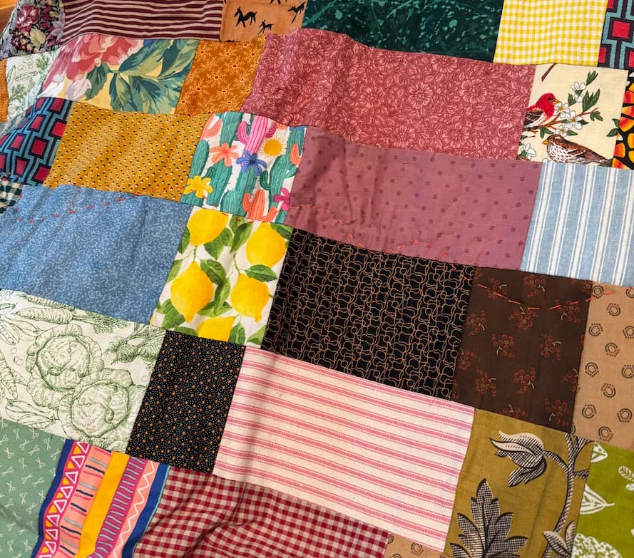 Colorful patchwork quilt or textile collage featuring diverse fabric patterns including florals, stripes, gingham, and solid colors in warm and cool tones.