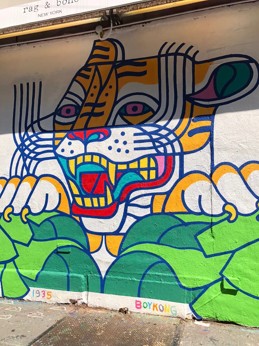 Colorful street mural by Boy Kong featuring a stylized face with bold geometric patterns in blue, yellow, orange, and green, with striped elements and expressive eyes.