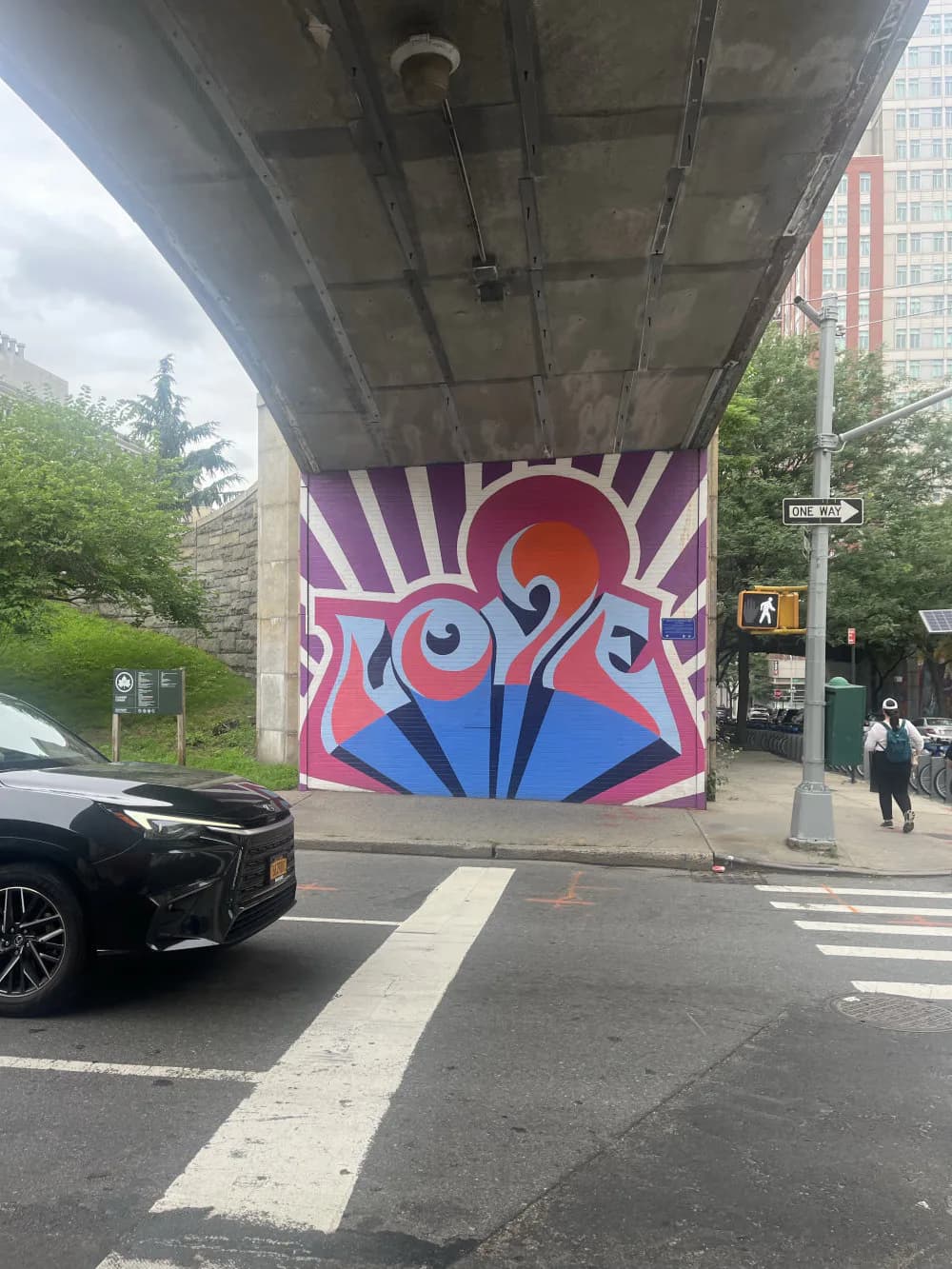 Large-scale mural under bridge underpass with pink, blue, and orange geometric abstract face design with radiating purple and white rays.