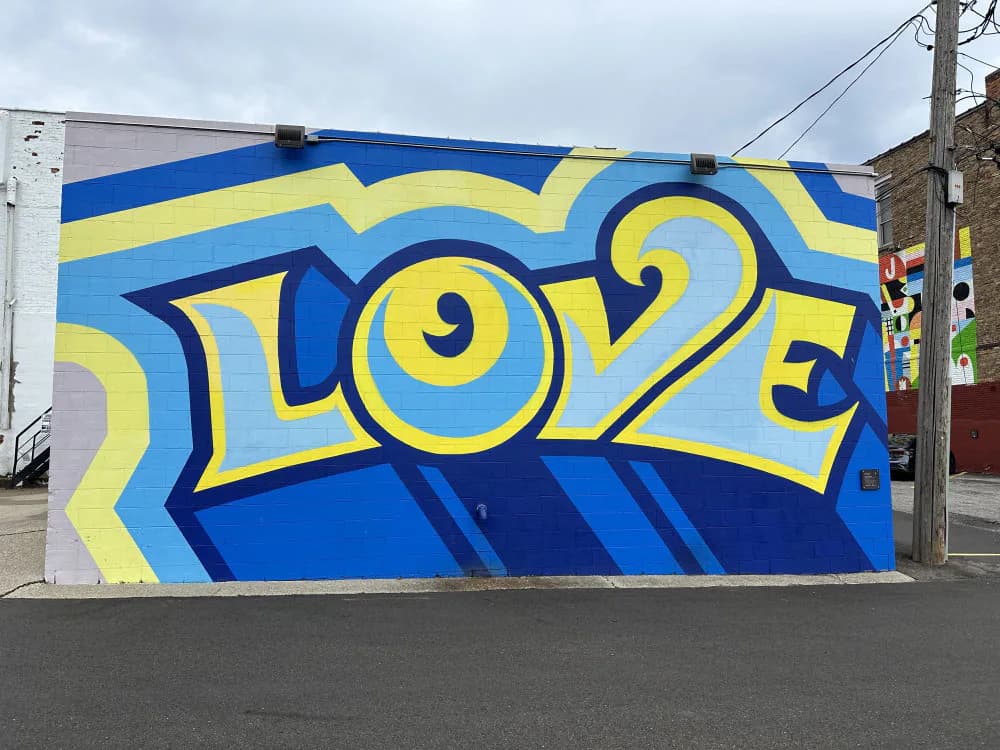 Mural featuring bold yellow and navy blue geometric lettering spelling 'SLOE2' with striped background and circular eye-like motifs.