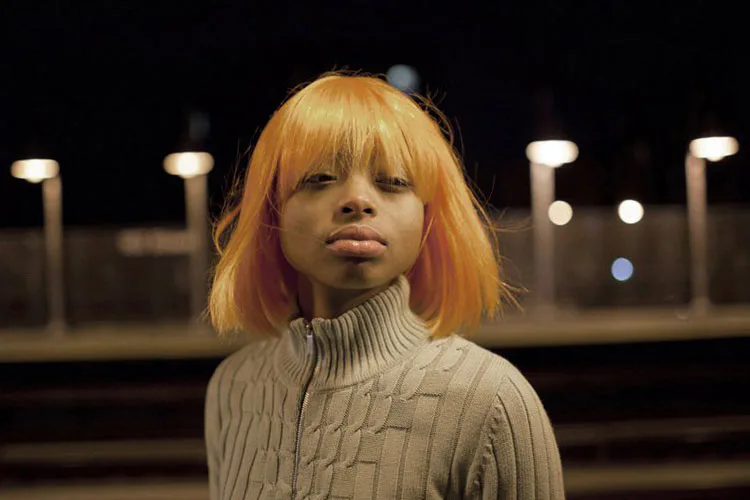 Young figure with bright orange bob haircut wearing cream cable-knit turtleneck, shot at night with blurred street lights in background.