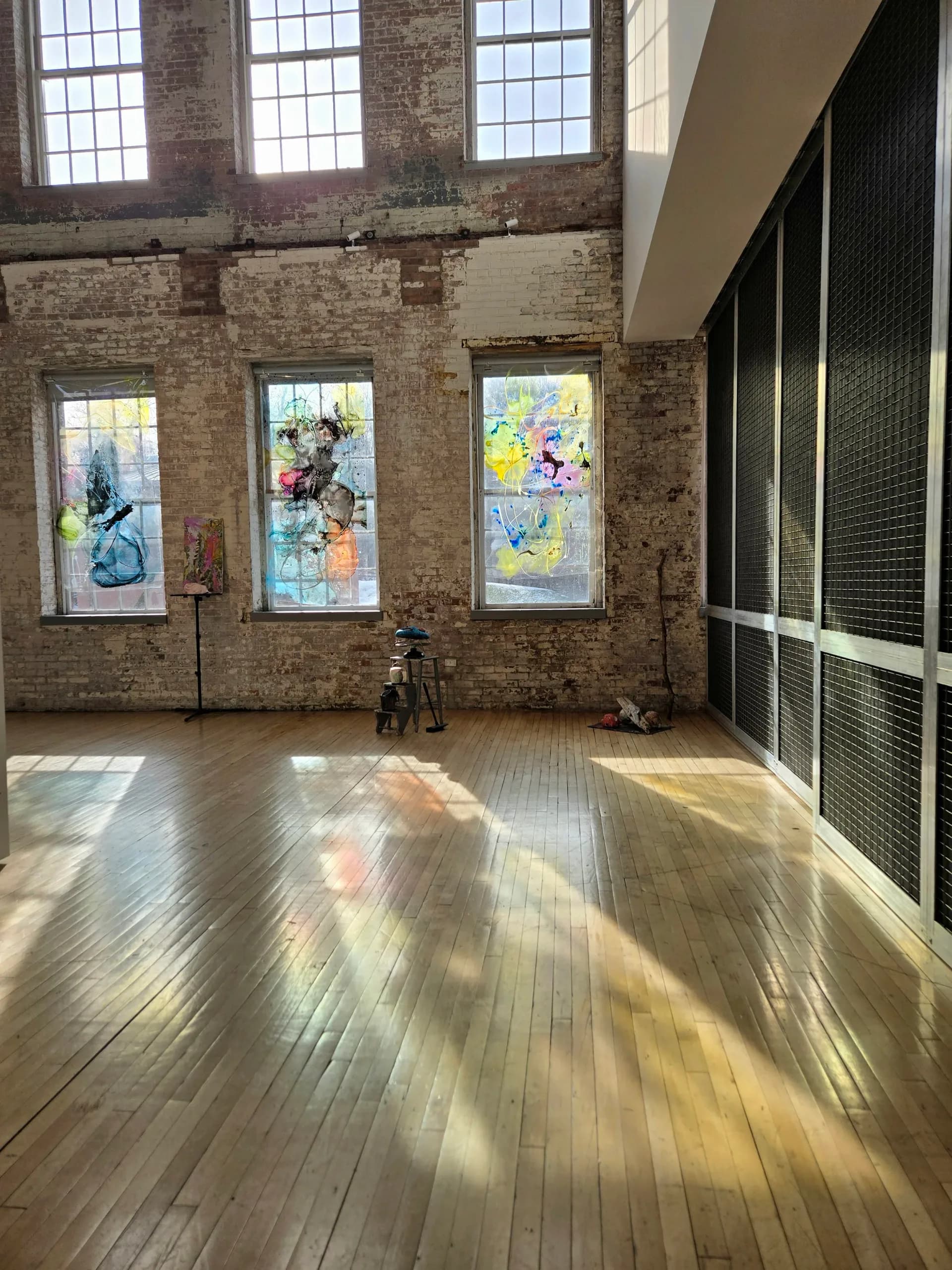 Interior gallery space with exposed brick walls, large windows, wooden floors; three framed artworks on walls featuring colorful abstract designs.