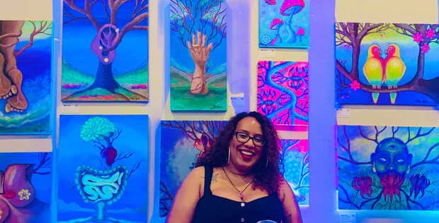 Woman wearing glasses smiling in front of a gallery wall displaying colorful neon-lit artworks featuring surreal faces and nature motifs.