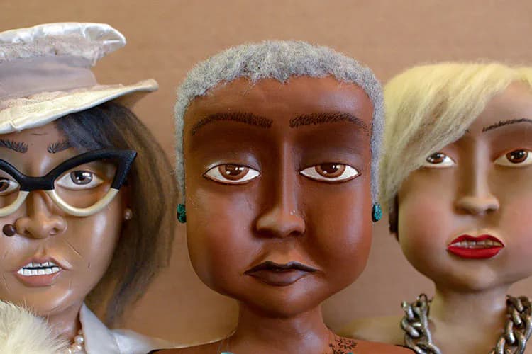 Three sculptural doll heads with diverse skin tones and styling: one wearing glasses and a hat, one with gray hair, one blonde. Polymer or resin sculpture.