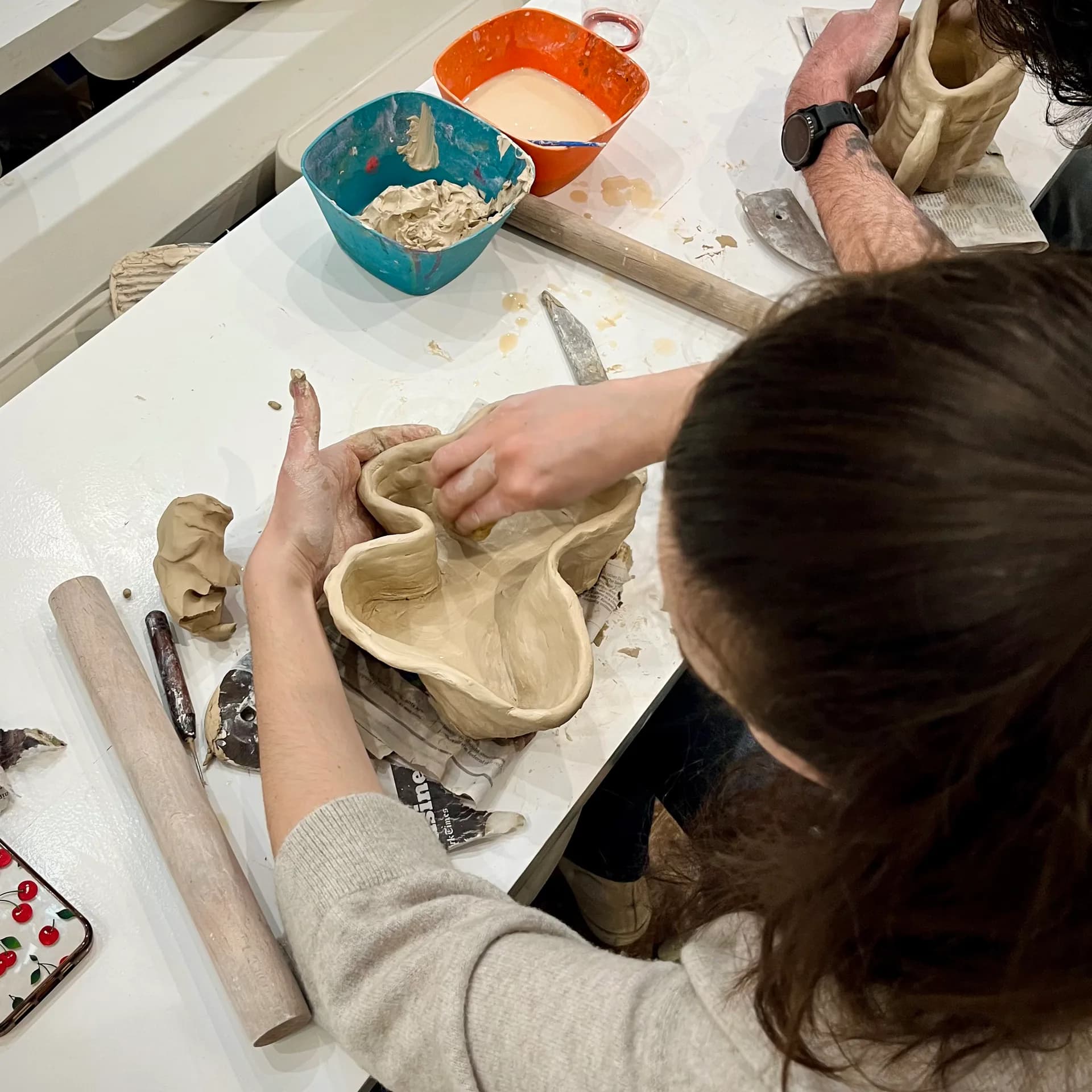 Student hand-building an organic clay vessel at a worktable with tools and slip