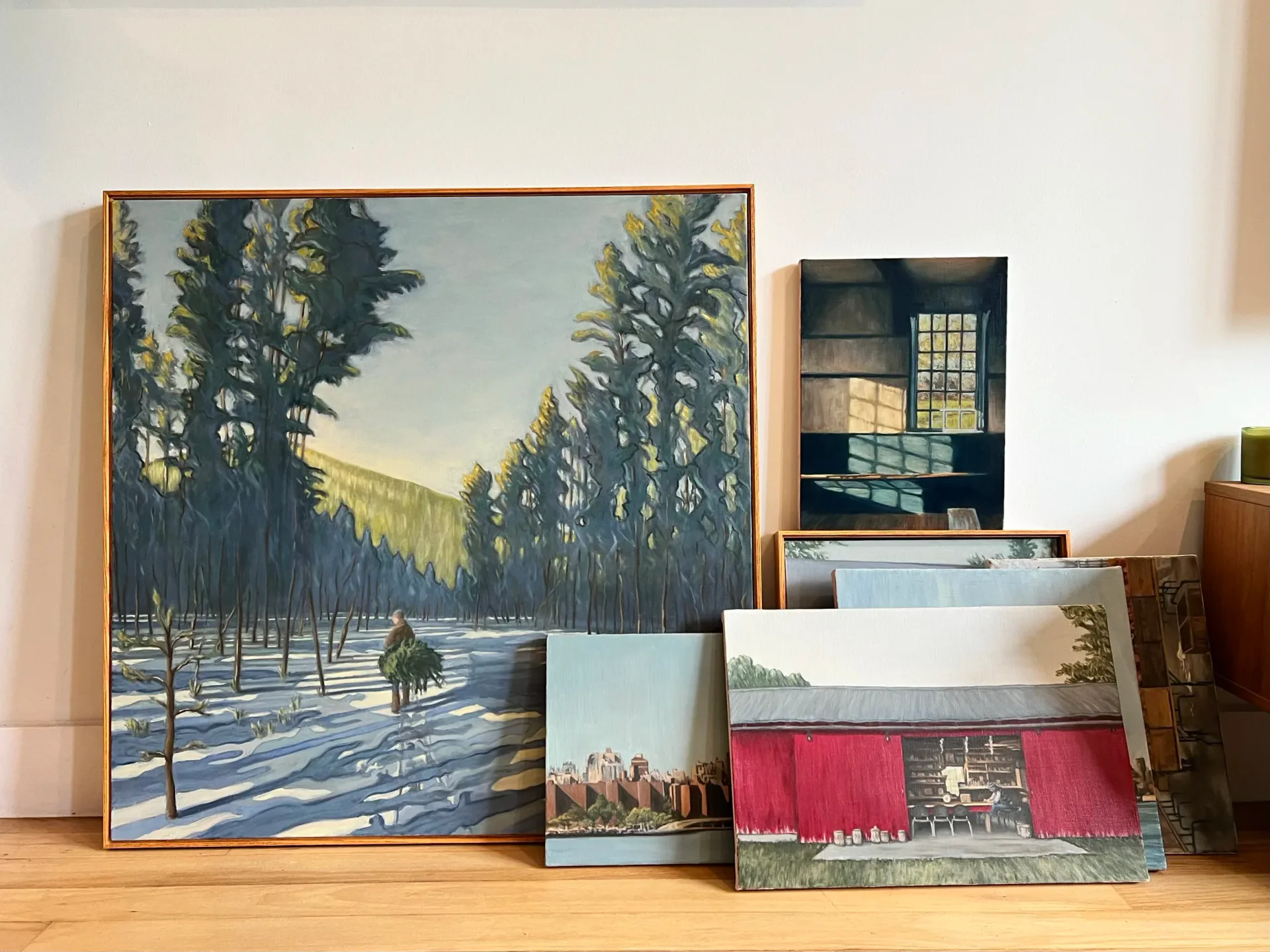 Photograph of framed paintings and artwork leaning against a wall in a studio or gallery space, including forest and architectural scenes by Cody Skinner.