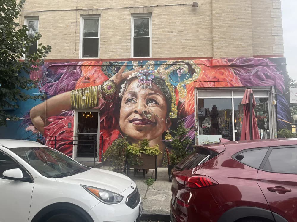 Vibrant mural by Damien Mitchell featuring a woman's portrait with purple, orange, and gold tones, adorned with jewelry and elaborate headdress against a floral background.