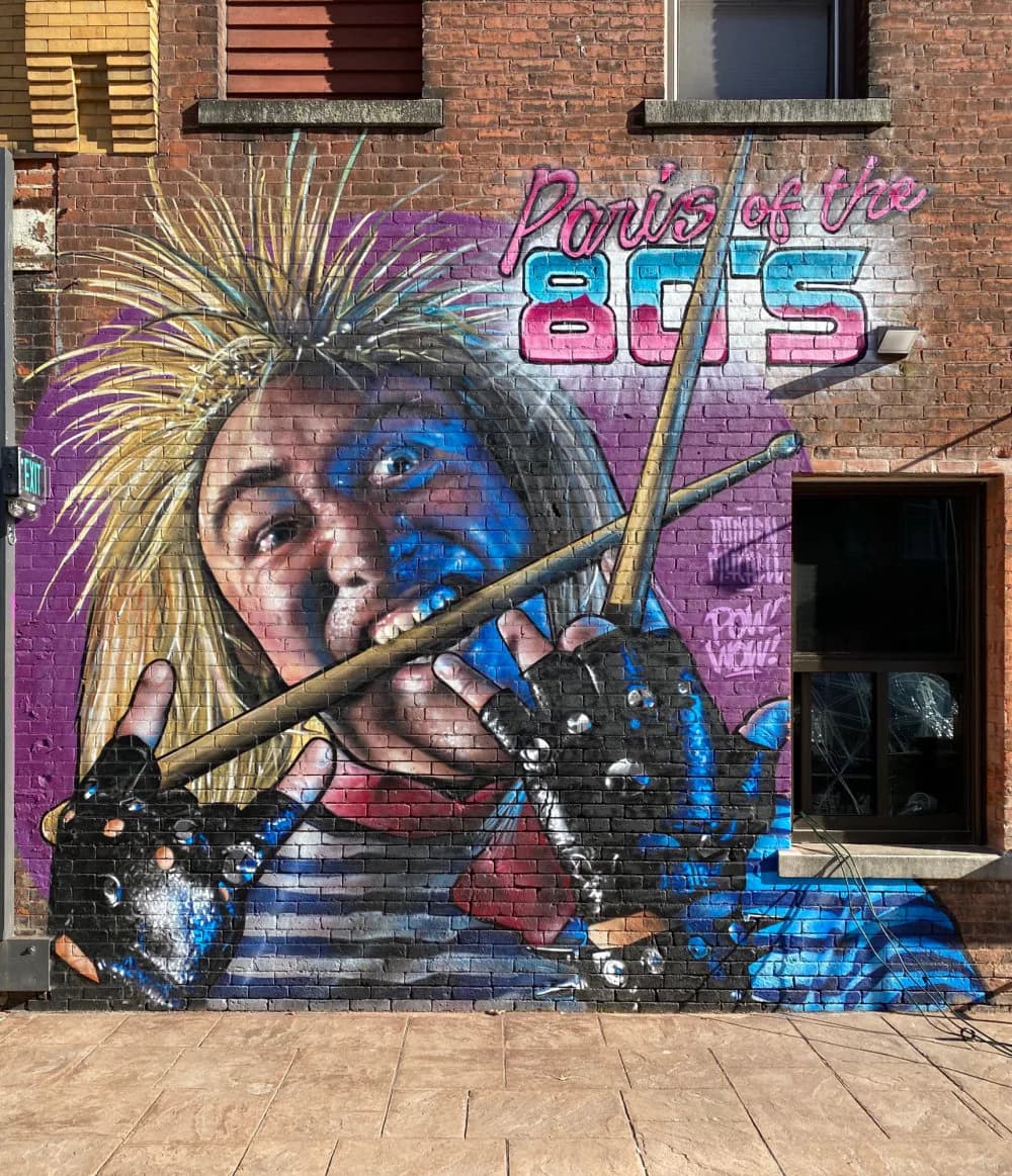 Mixed-media mural combining portrait photography, blue geometric shapes, purple typography reading 'Points of View 80s', and yellow starburst elements on brick.