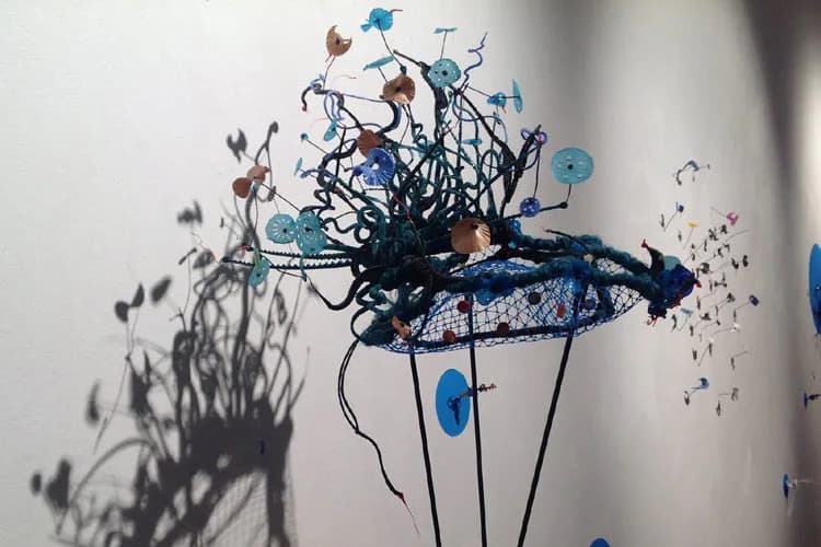 Abstract mixed-media sculpture featuring tangled black wire and mesh forms with blue circular elements and scattered particles, casting dramatic shadows on white wall.