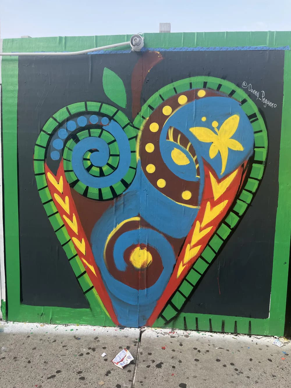 Vibrant mural by Danny Peguero depicting a stylized heart with blue, red, yellow, and green geometric patterns, spirals, and flame motifs on a dark background.