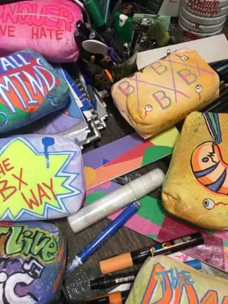 Flat-lay arrangement of colorful fabric pouches and markers with bold graffiti-style text and designs in yellow, pink, blue, and purple.