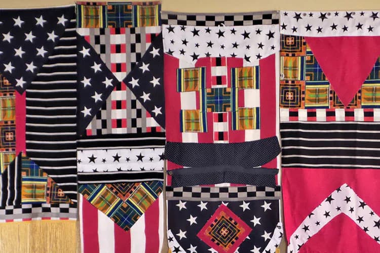 Textile quilt with patchwork panels combining red, navy, and white fabrics featuring stars, stripes, and geometric patterns with gold/yellow accents.