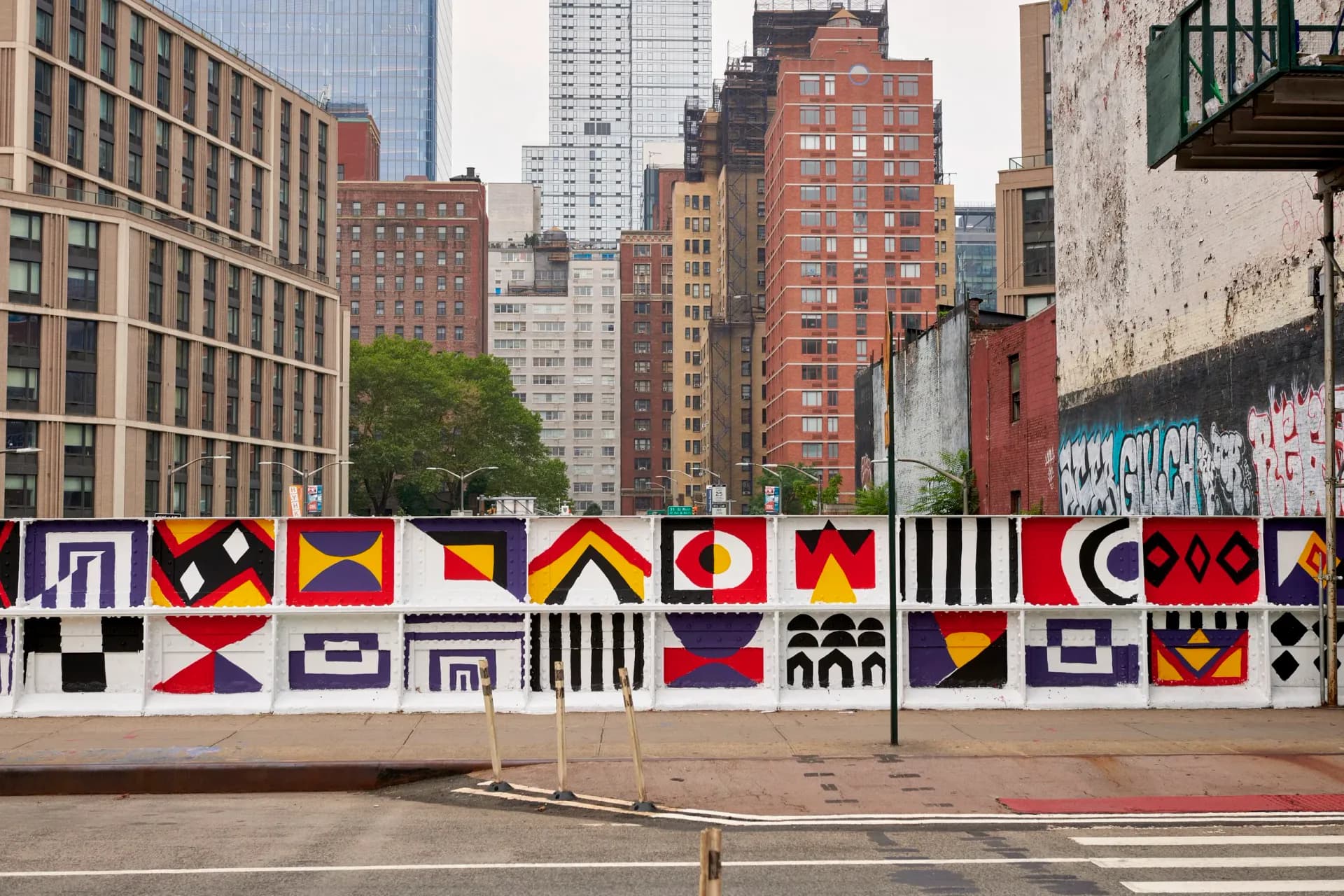 Large-scale outdoor mural on urban wall featuring geometric patterns in red, blue, yellow, black, and white with abstract diamond and linear designs.