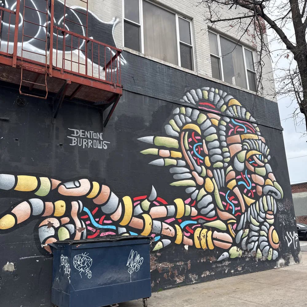 Mural on dark background featuring a hand with intricate mechanical and organic details in yellow, red, and blue by artist Denton Burrows.