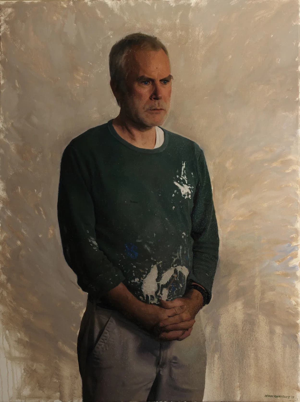 Man in dark green long-sleeve shirt with graphic paint splatters, hands clasped, standing against textured beige wall.