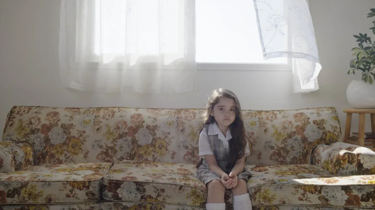 Young girl in school uniform sitting alone on floral sofa with billowing curtain