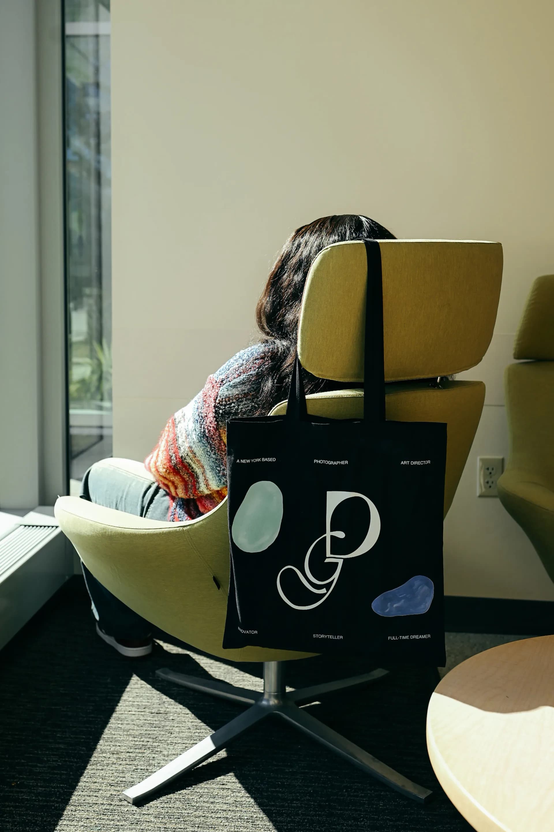 Person seated in mustard chair holding black tote bag with white abstract line drawings and logos.