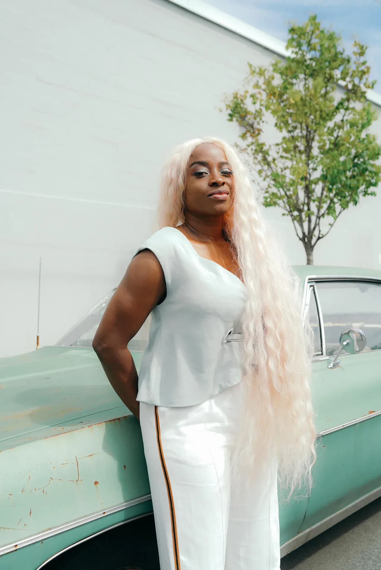Woman in white sleeveless outfit with long white hair posing against mint-green vintage car.