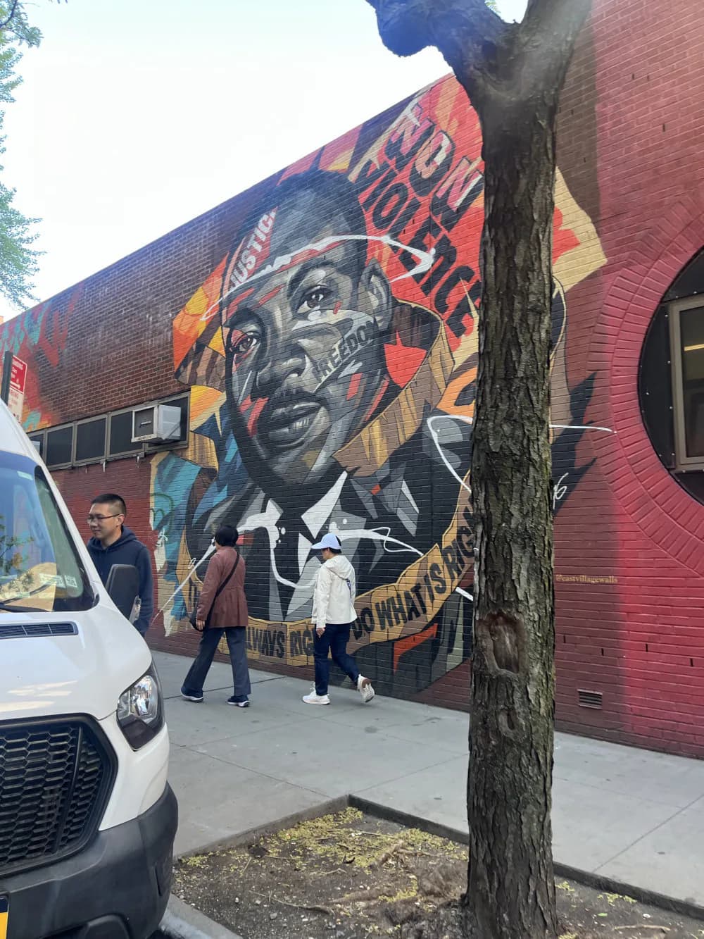Street mural on brick building showing detailed portrait with geometric overlays in red, orange, and blue; people stand before it for scale.