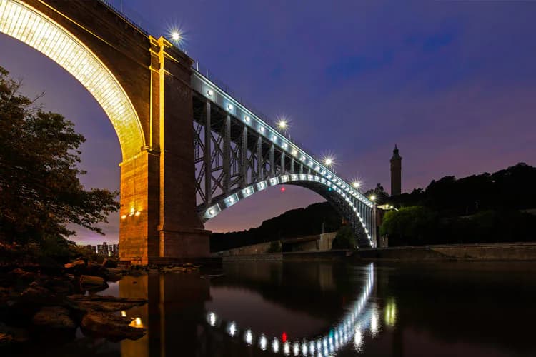 Illuminated arch bridge spanning a river at dusk, reflected in calm water below, with a tower visible in the background and purple twilight sky.