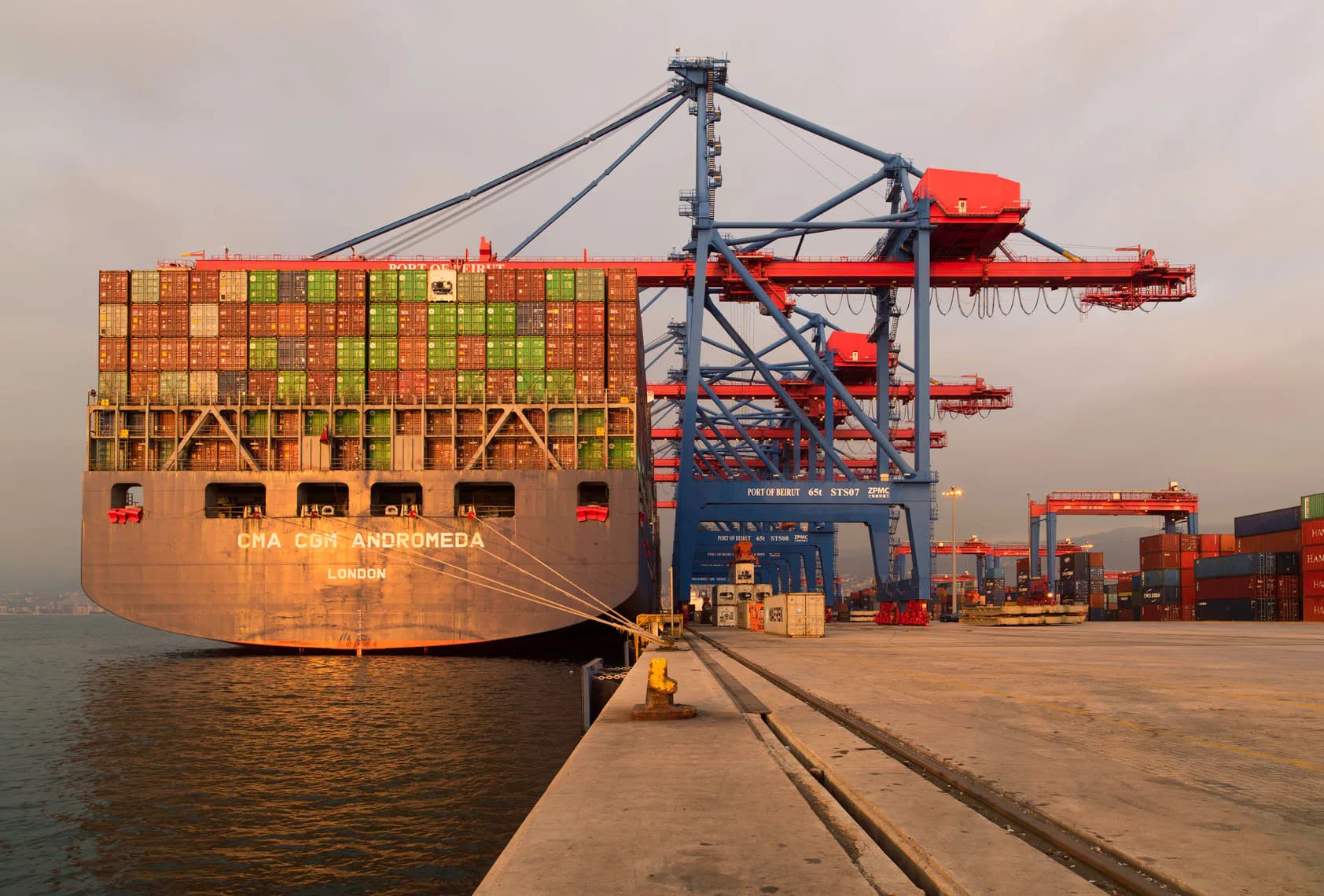 Container ship loaded with stacked cargo boxes docked at industrial port with red cranes, shot at golden hour.