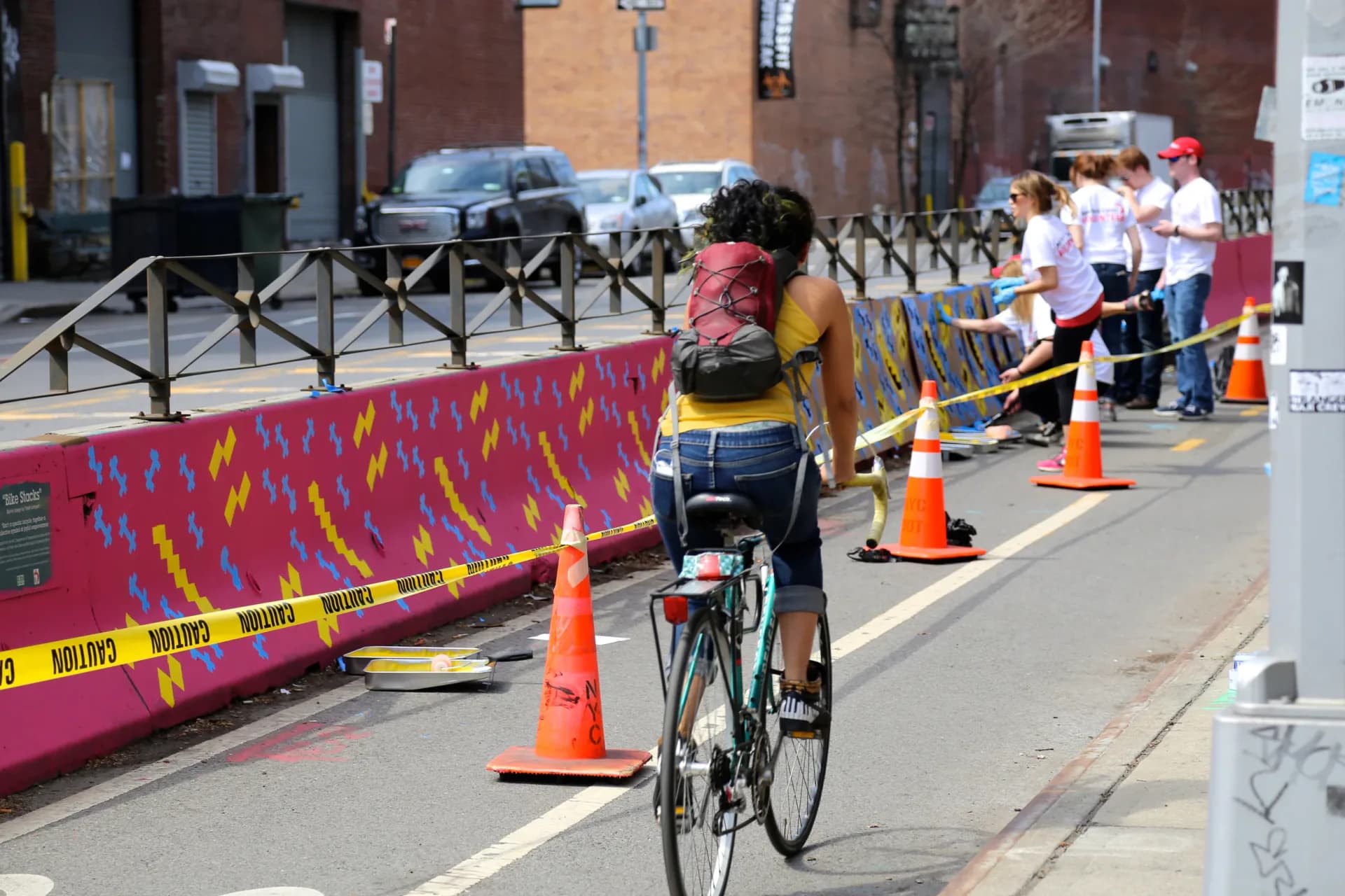 Outdoor street scene with a cyclist at a community event, traffic cones, colorful barrier wall, and spectators in background.