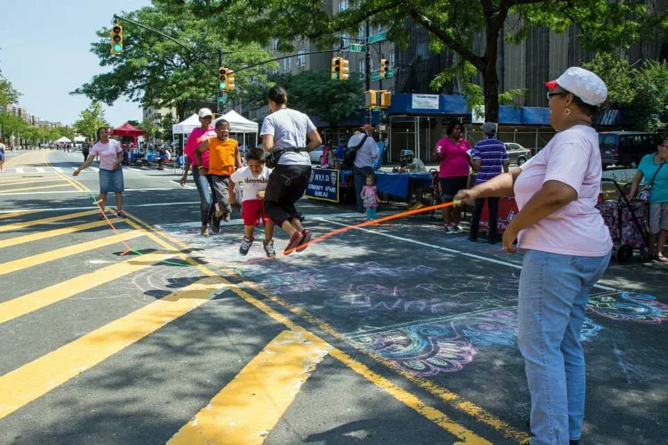 Outdoor community gathering on painted street with yellow lines, people jumping rope, trees, and urban buildings visible.