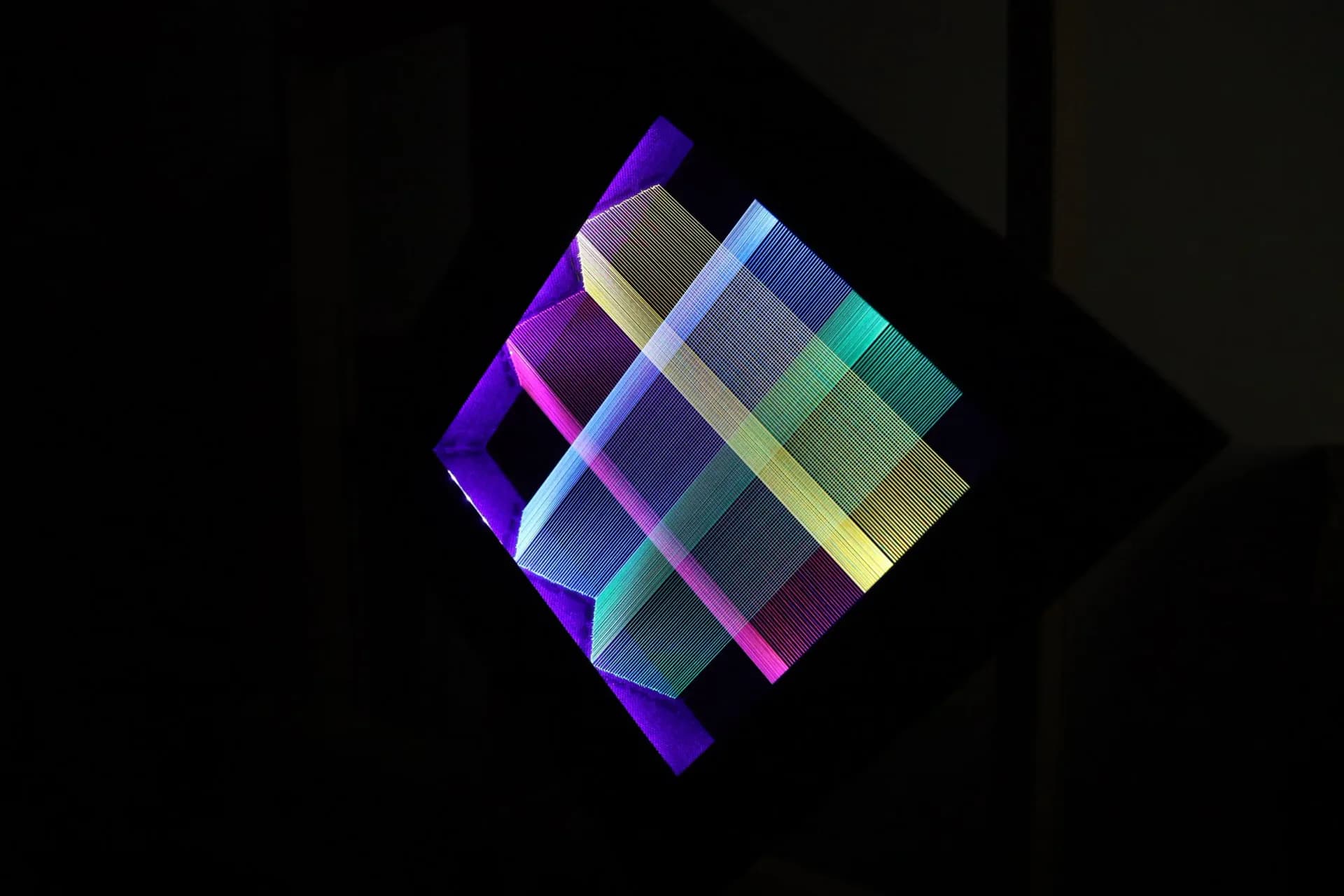 Diamond-shaped framed string art with intersecting blue, teal, yellow and pink threads glowing against black