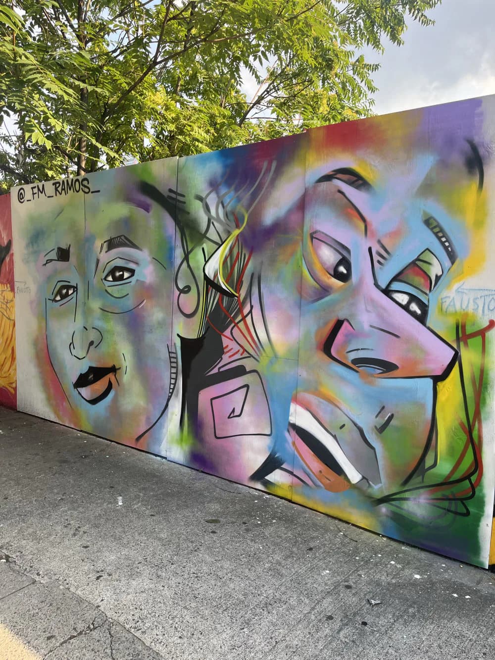 Colorful mural by Fausto Manuel Ramos featuring three expressive faces with rainbow gradients, bold outlines, and mixed-media style on concrete wall.