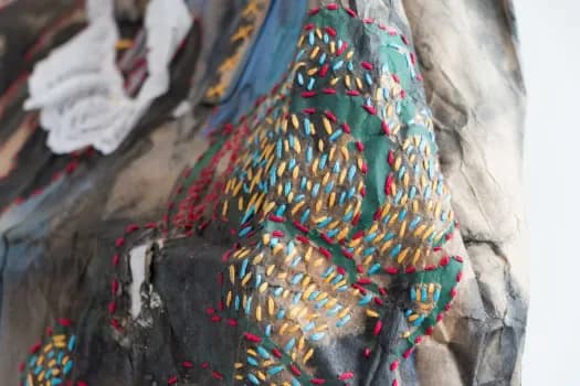 Close-up of mixed-media textile artwork with embroidered or stitched multicolored geometric patterns (orange, green, red, yellow) applied over a painted fabric base in blue and gray tones.